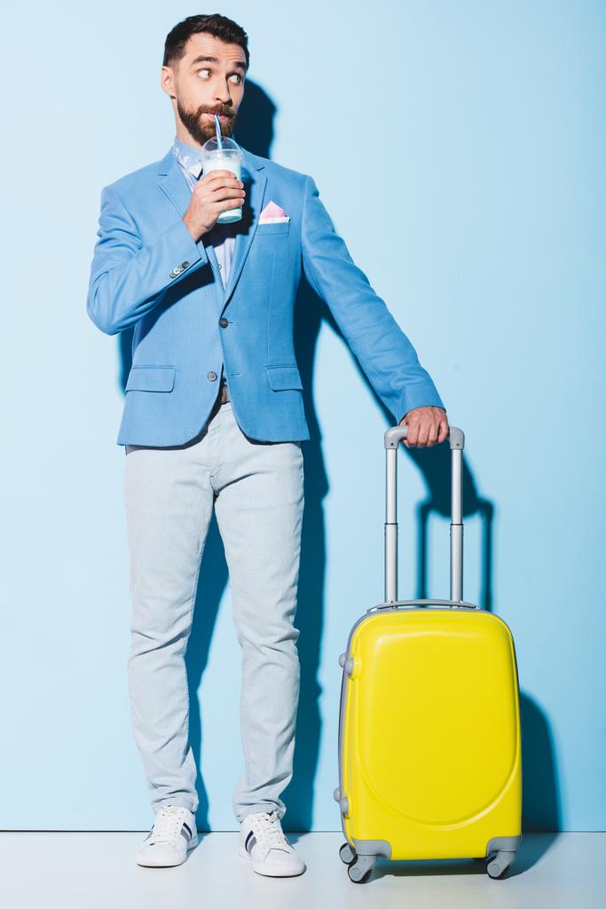 άνθρωπος πίνοντας κοκτέιλ και κρατώντας τσάντα ταξιδιού σε μπλε φόντο  - Φωτογραφία, εικόνα