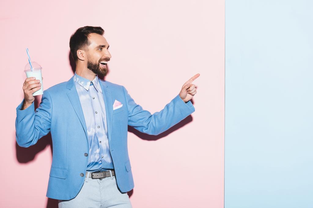  χαμογελαστός άνδρας κρατώντας κοκτέιλ και δείχνοντας με το δάχτυλο σε ροζ και μπλε φόντο  - Φωτογραφία, εικόνα