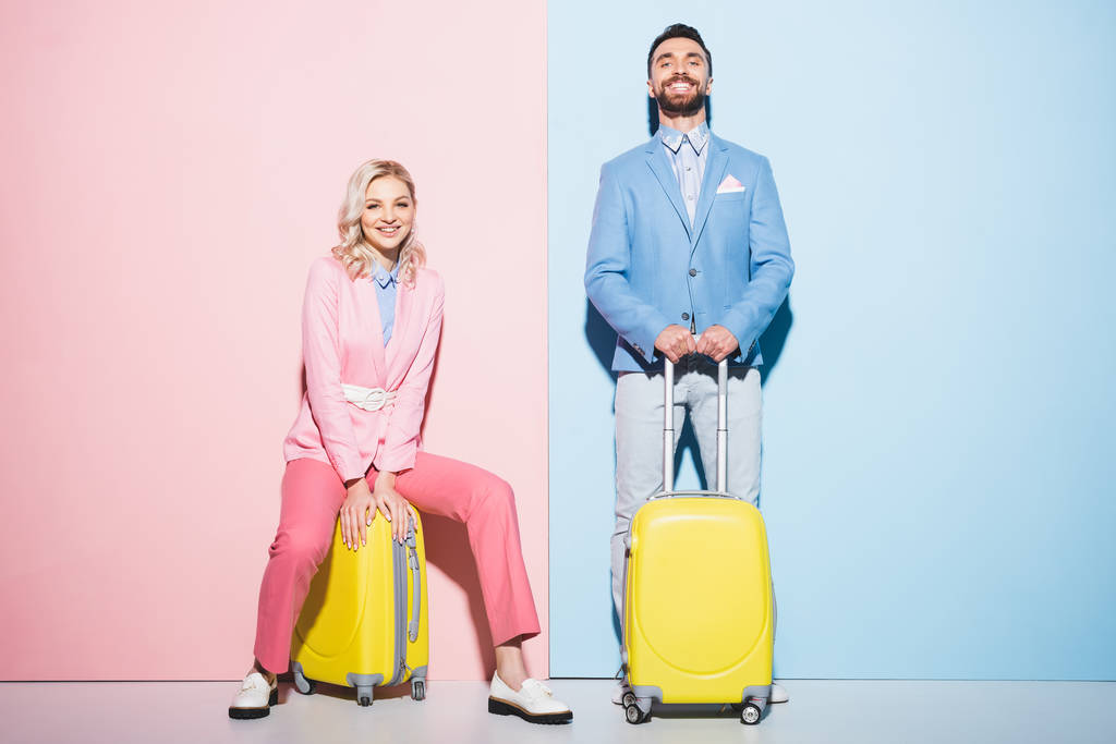 glimlachende vrouw en knappe man zitten op reistassen op roze en blauwe achtergrond  - Foto, afbeelding