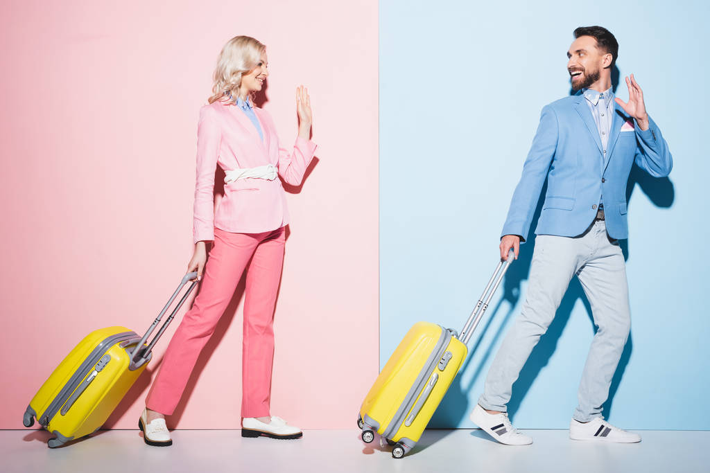 uśmiechnięta kobieta i przystojny mężczyzna z torbami podróżnymi machając na różowym i niebieskim tle  - Zdjęcie, obraz