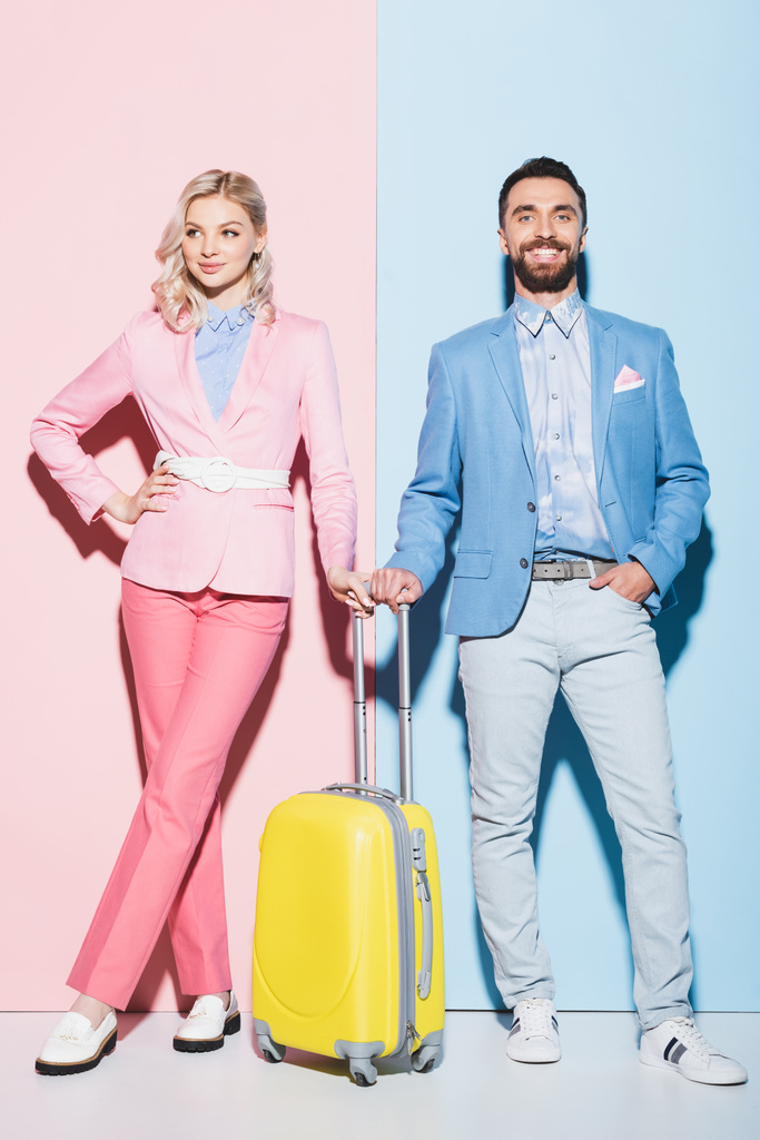 χαμογελαστή γυναίκα και όμορφος άντρας με ταξιδιωτική τσάντα σε ροζ και μπλε φόντο  - Φωτογραφία, εικόνα