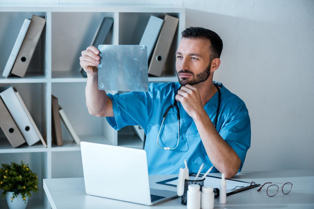 ortopedista guapo mirando rayos X cerca del ordenador portátil en la clínica
  - Foto, Imagen