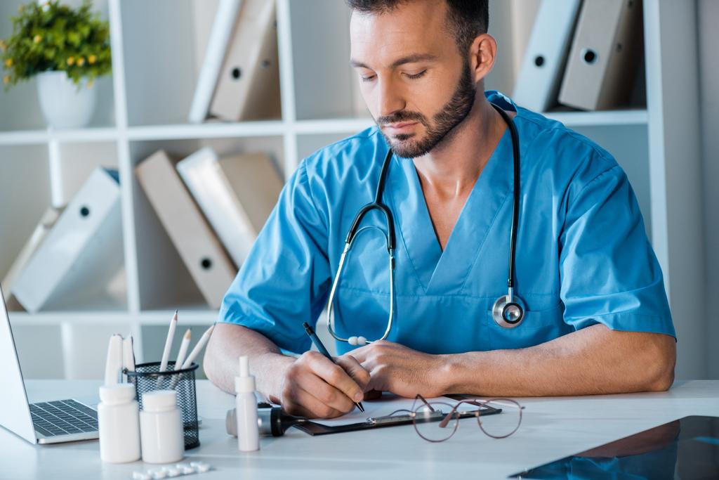 handsome doctor writing prescription near laptop in clinic  - Photo, Image