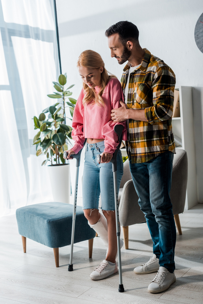 handsome bearded man standing with injured woman holding crutches at home  - Photo, Image
