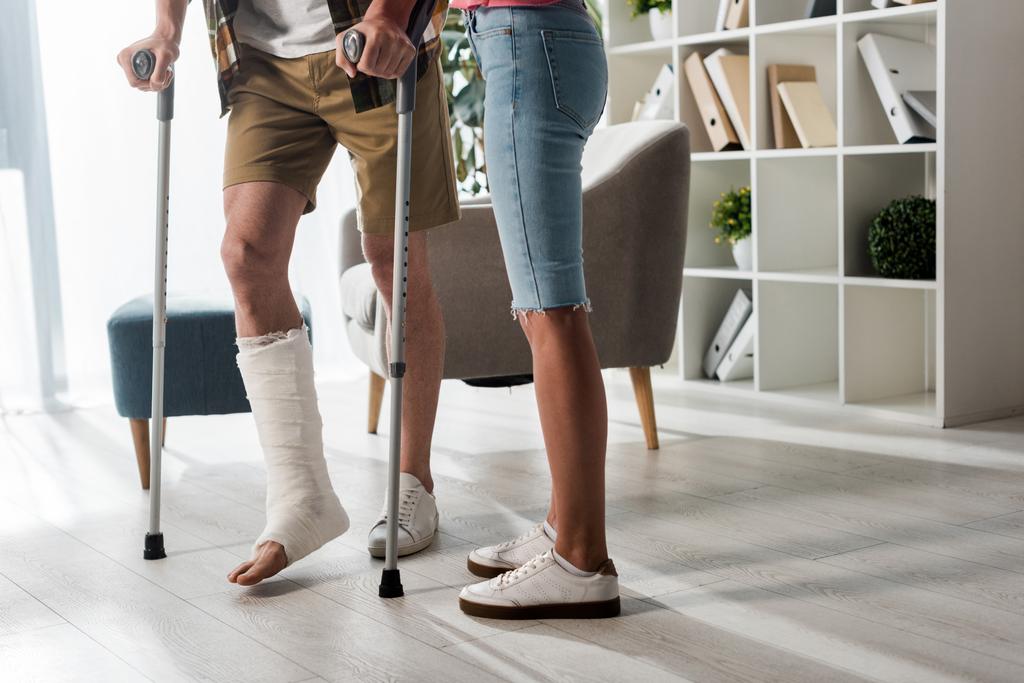 cropped view of woman standing near man holding crutches at home  - Fotoğraf, Görsel