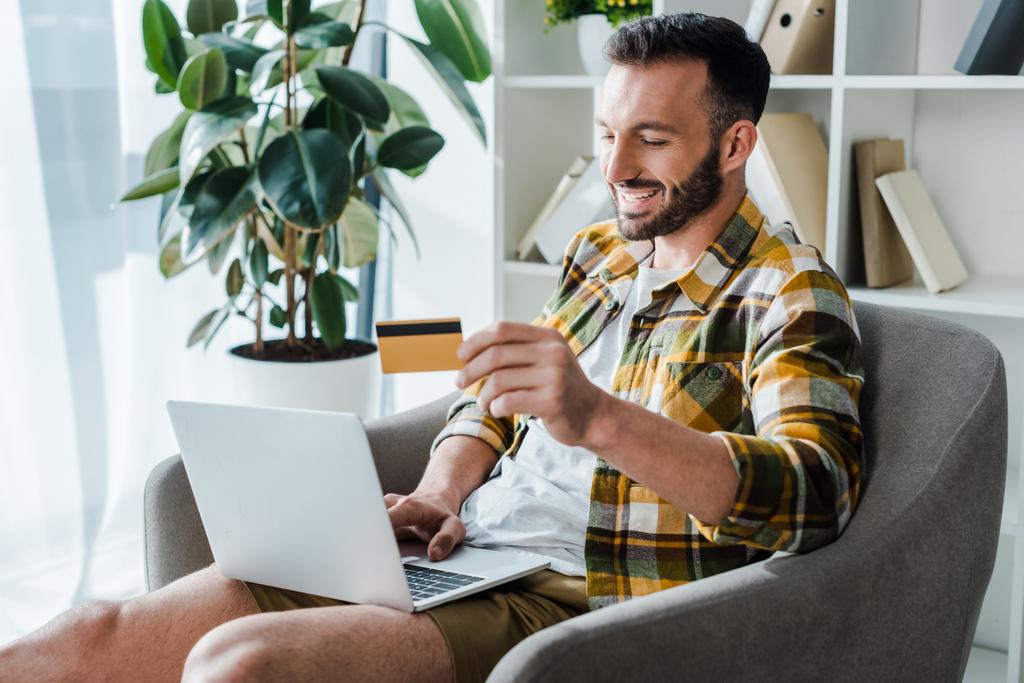handsome and smiling man holding credit card while online shopping at home  - Fotoğraf, Görsel