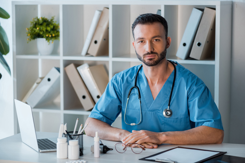 pohledný vousatý lékař sedí u brýlí a notebooku na klinice  - Fotografie, Obrázek