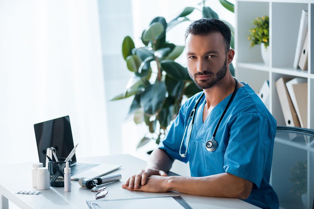 schöner bärtiger Arzt blickt in der Nähe von Laptop in Klinik in die Kamera  - Foto, Bild