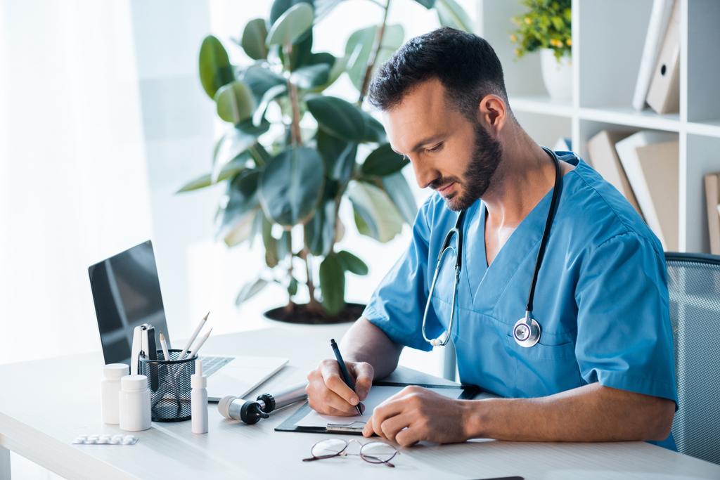 przystojny brodaty lekarz piszący diagnozę w klinice  - Zdjęcie, obraz