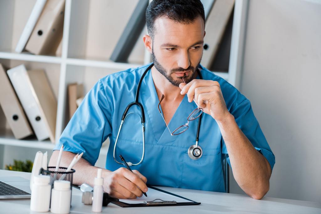 Nachdenklicher Arzt hält Stift in der Nähe von Klemmbrett in Klinik  - Foto, Bild
