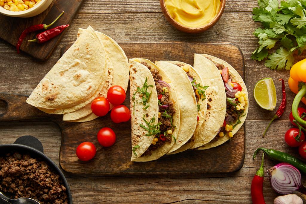 Bovenaanzicht van smakelijke taco 's op snijplank met ingrediënten op houten achtergrond - Foto, afbeelding