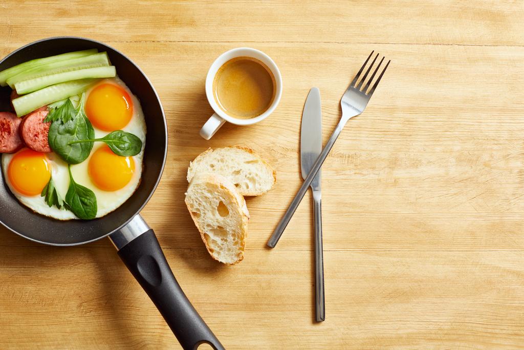 πάνω όψη τηγανιτών αυγών με φύλλα σπανάκι, αγγούρι και λουκάνικο σε τηγάνι κοντά σε καφέ, ψωμί και καφέ σε ξύλινο τραπέζι - Φωτογραφία, εικόνα