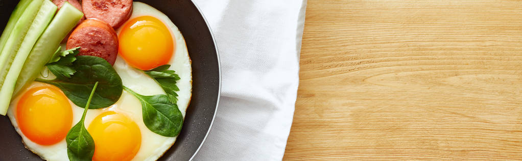 felülnézet sült tojás spenótlevéllel, uborkával és kolbásszal serpenyőben a szalvétán a fa asztalon, panorámás felvétel - Fotó, kép