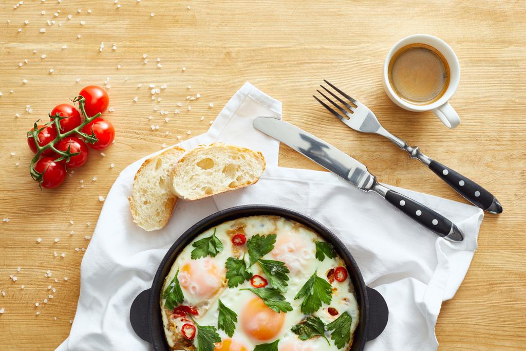 Tavada tavada yumurta, çatal bıçak, kahve ve domatesler ahşap masada. - Fotoğraf, Görsel
