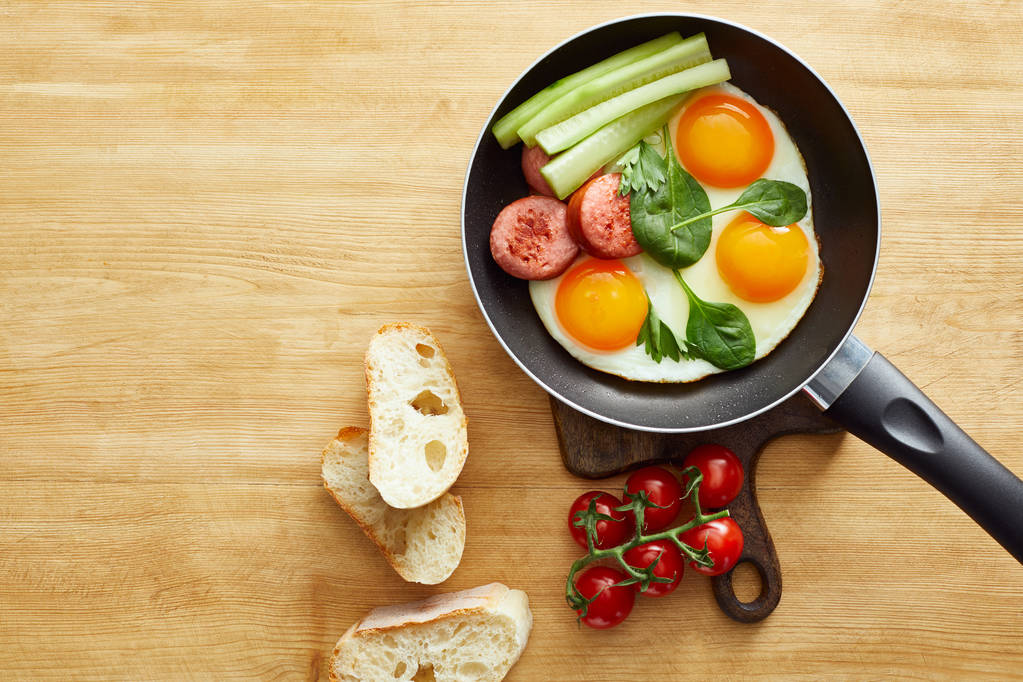 vista superior de ovos fritos com folhas de espinafre, pepino e salsicha na frigideira perto de tomates e pão na mesa de madeira
 - Foto, Imagem