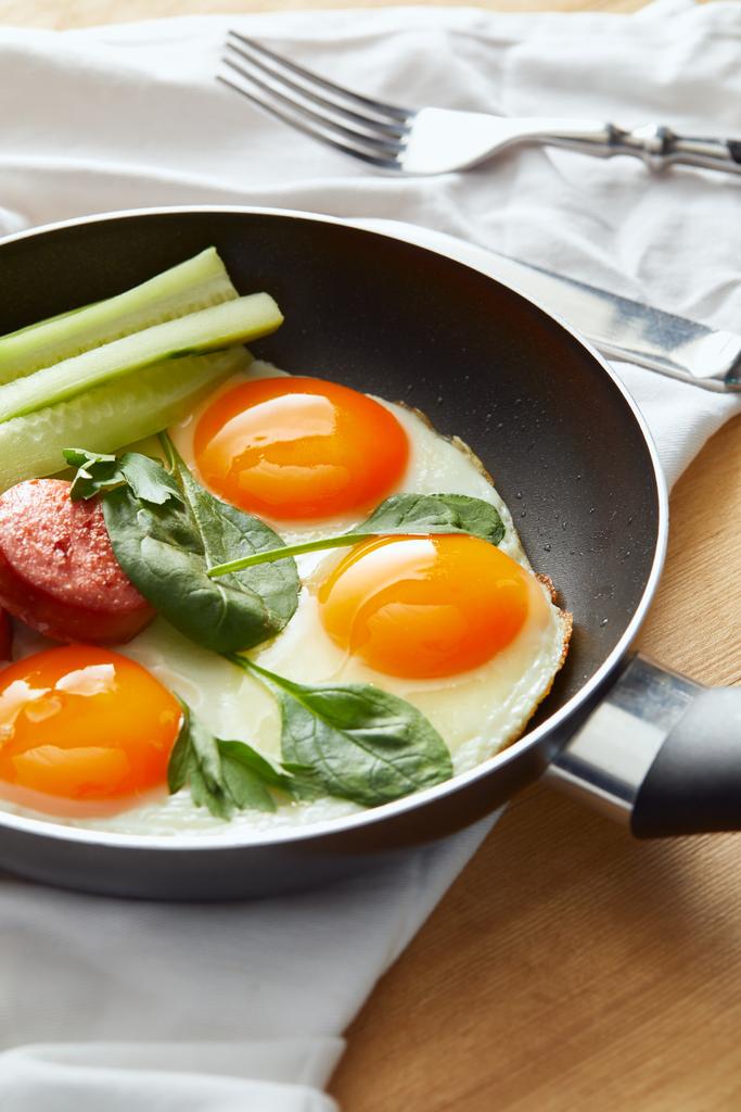 selective focus of fried eggs in frying pan with spinach, cucumber and sausage at wooden table with cutlery on napkin - Photo, Image