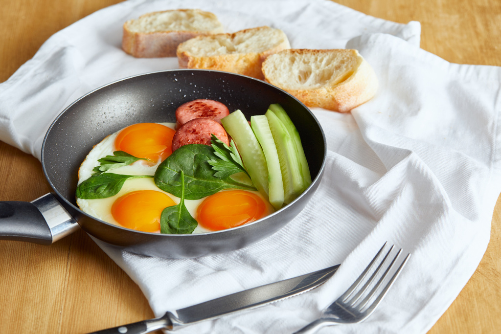 селективный фокус яиц на сковороде со шпинатом, огурцом и колбасой за деревянным столом со столовыми приборами и хлебом на салфетке
 - Фото, изображение
