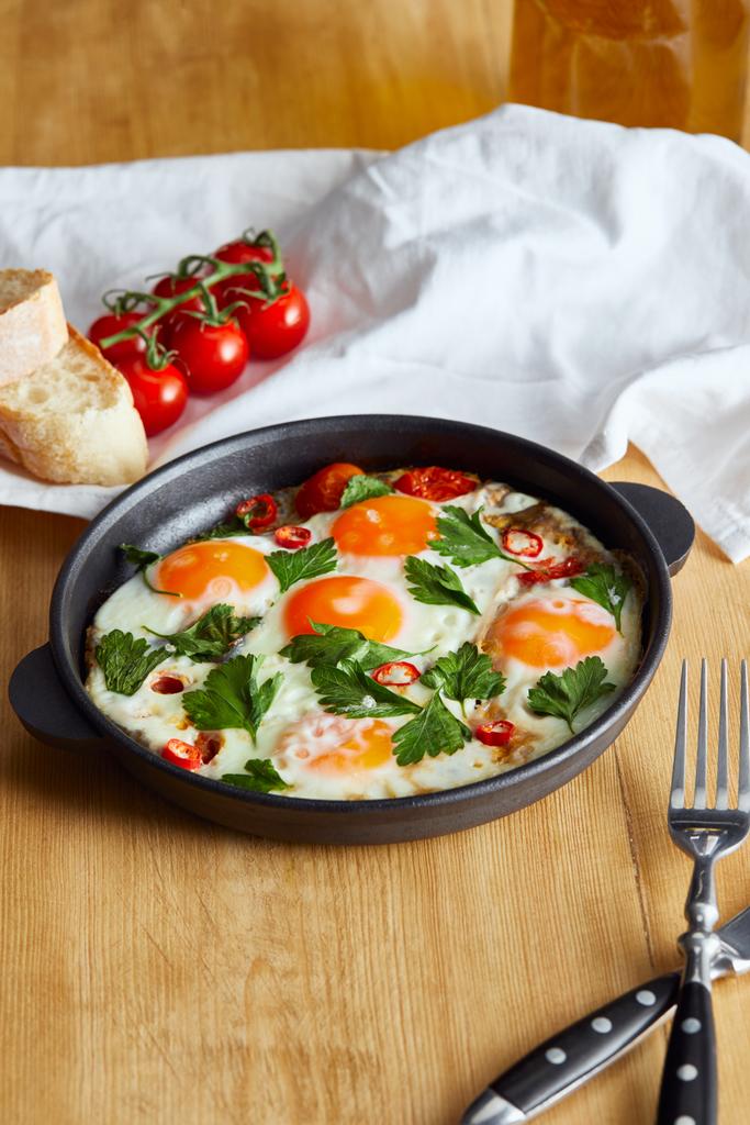 fried eggs with parsley and chili pepper in pan near napkin, bread, tomatoes and napkin on wooden table - Photo, Image