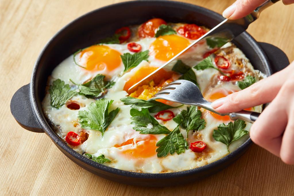 cropped view of eating fried eggs with parsley and chili pepper on wooden table with fork and knife - Photo, Image