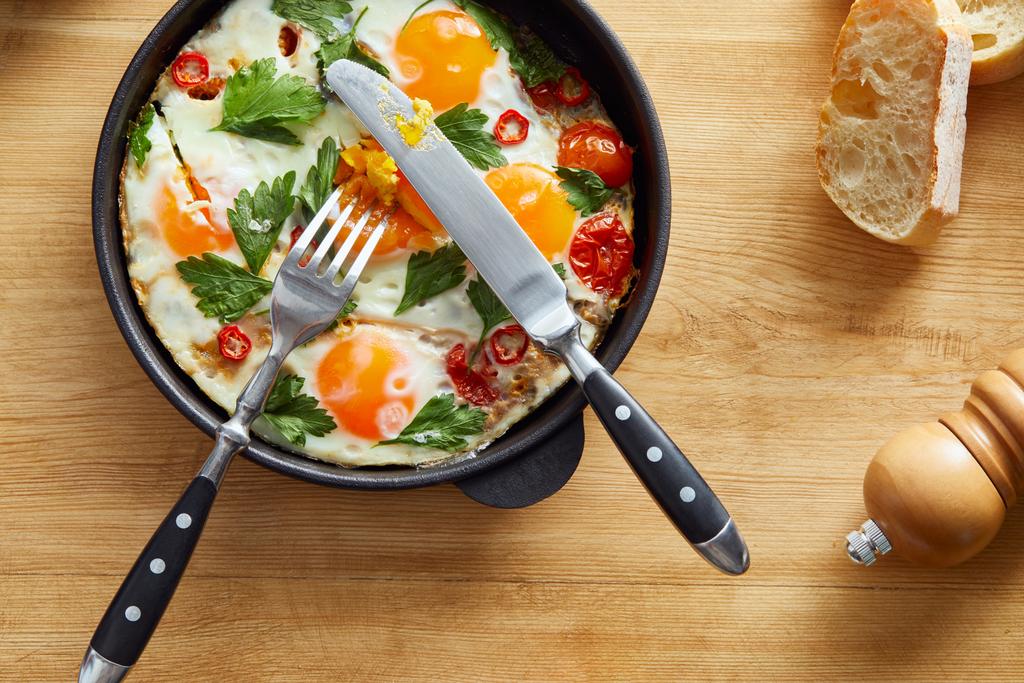 top view of fried eggs with parsley and chili pepper on wooden table with cutlery and bread - Photo, Image