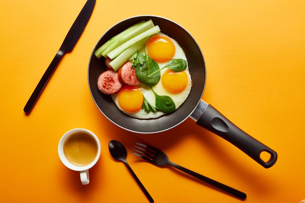 vrchní pohled na smažená vejce se špenátovými listy, okurkou a klobásou na pánvi u kávy, příbory na oranžovém pozadí - Fotografie, Obrázek