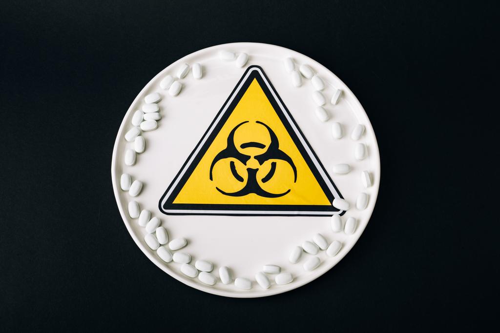 Κάτοψη του πιάτου με τα χάπια και το σύμβολο βιολογικού κινδύνου που απομονώνονται σε μαύρο - Φωτογραφία, εικόνα