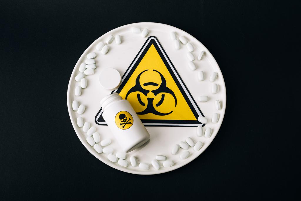 Bovenaanzicht van biologisch gevarenbord op bord met pillen en fles met giftige letters geïsoleerd op zwart - Foto, afbeelding
