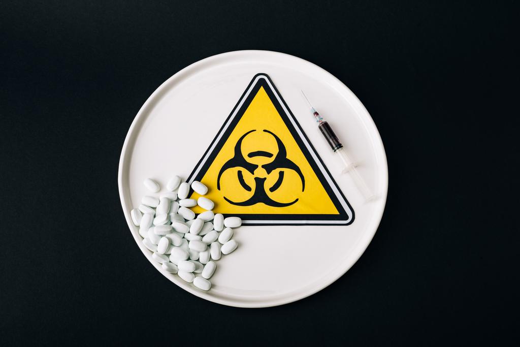 Vista superior de píldoras y jeringa en placa con signo de riesgo biológico aislado en negro
 - Foto, Imagen