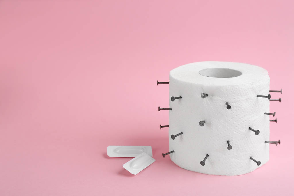 Рулон туалетной бумаги с гвоздями и свечами на розовом фоне, место для текста. Геморрой проблемы
 - Фото, изображение