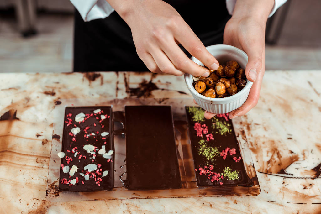 bijgesneden weergave van chocolatier holding bowl met gekarameliseerde hazelnoten in de buurt van chocoladerepen  - Foto, afbeelding