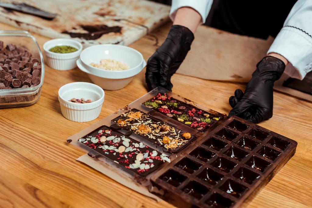 обрезанный вид шоколада в черных латексных перчатках, держащих шоколадные формы с готовыми шоколадными батончиками
  - Фото, изображение