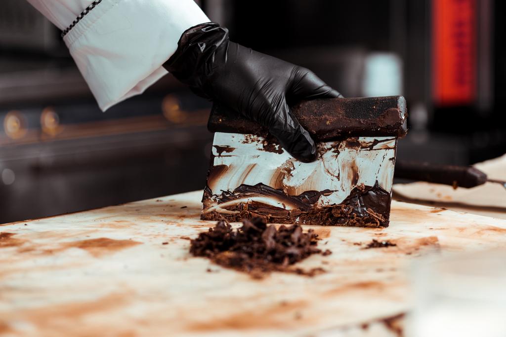 Ausgeschnittene Ansicht eines Chocolatiers in schwarzem Latex-Handschuh, der dunkle Schokolade von der Oberfläche kratzt  - Foto, Bild
