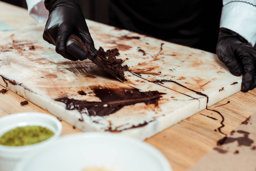 bijgesneden weergave van chocolatier in zwarte latex handschoenen schrapen pure chocolade krullen  - Foto, afbeelding