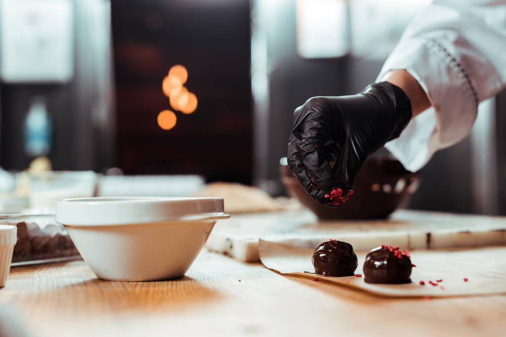 bijgesneden weergave van chocolatier in zwarte latex handschoen toevoegen van gedroogde frambozen op vers gemaakte snoepjes  - Foto, afbeelding
