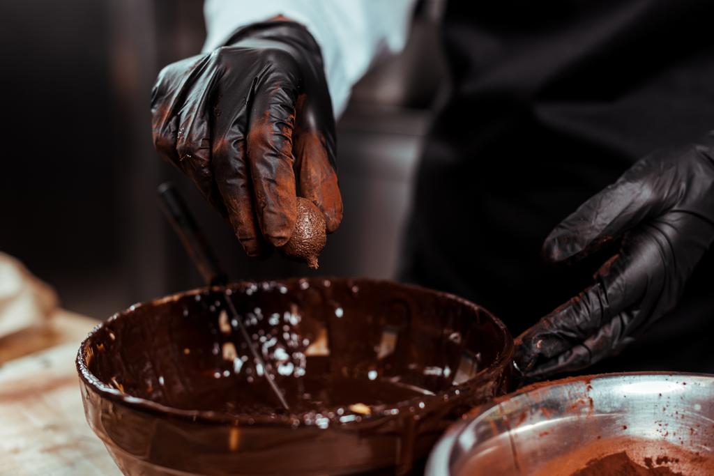vista cortada de chocolate em luva de látex preto segurando bola de chocolate perto da tigela com chocolate derretido
  - Foto, Imagem