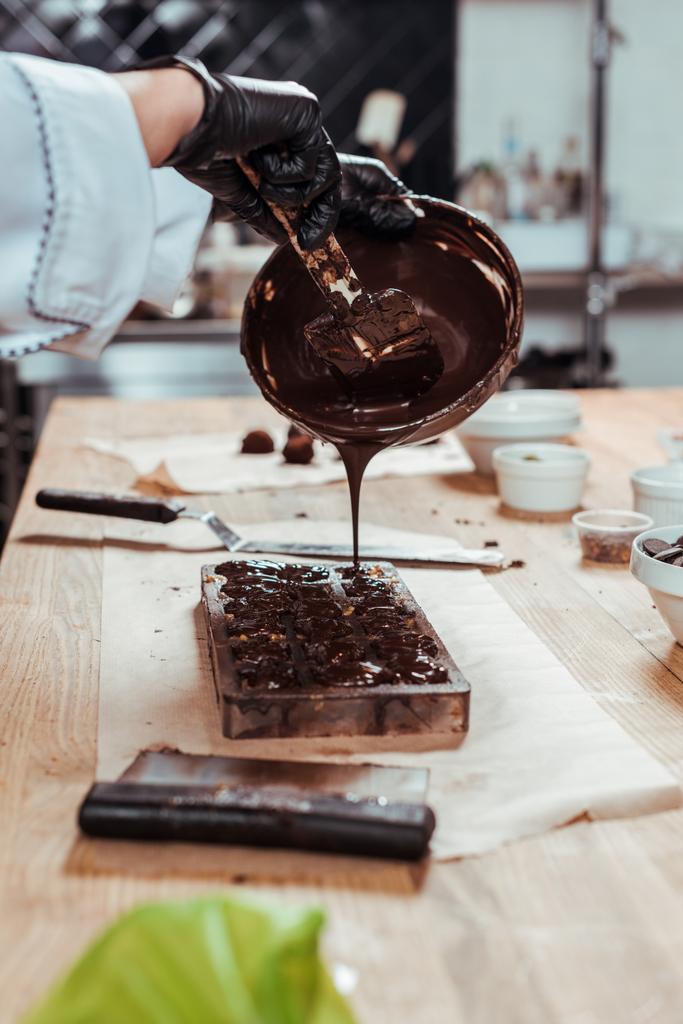 cropped άποψη της σοκολάτας κρατώντας μπολ, ενώ ρίχνει λιωμένη σοκολάτα σε καλούπια σοκολάτας στο τραπέζι  - Φωτογραφία, εικόνα