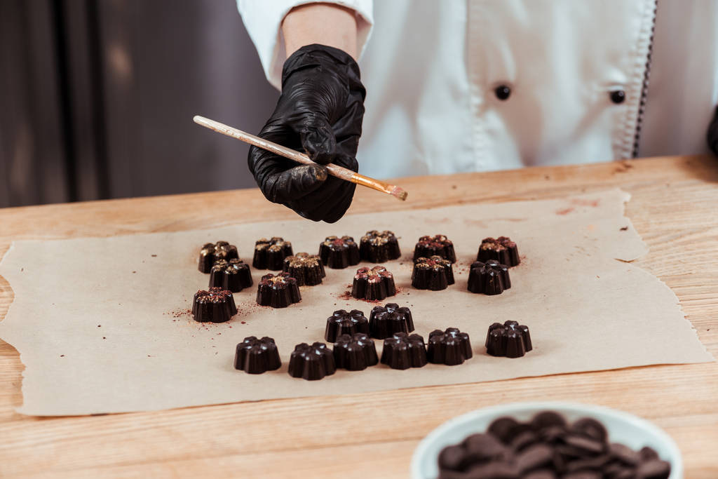 обрізаний вид шоколатьє, що тримає пензлик із золотим порошком біля смачних шоколадних цукерок на папері для випічки
  - Фото, зображення