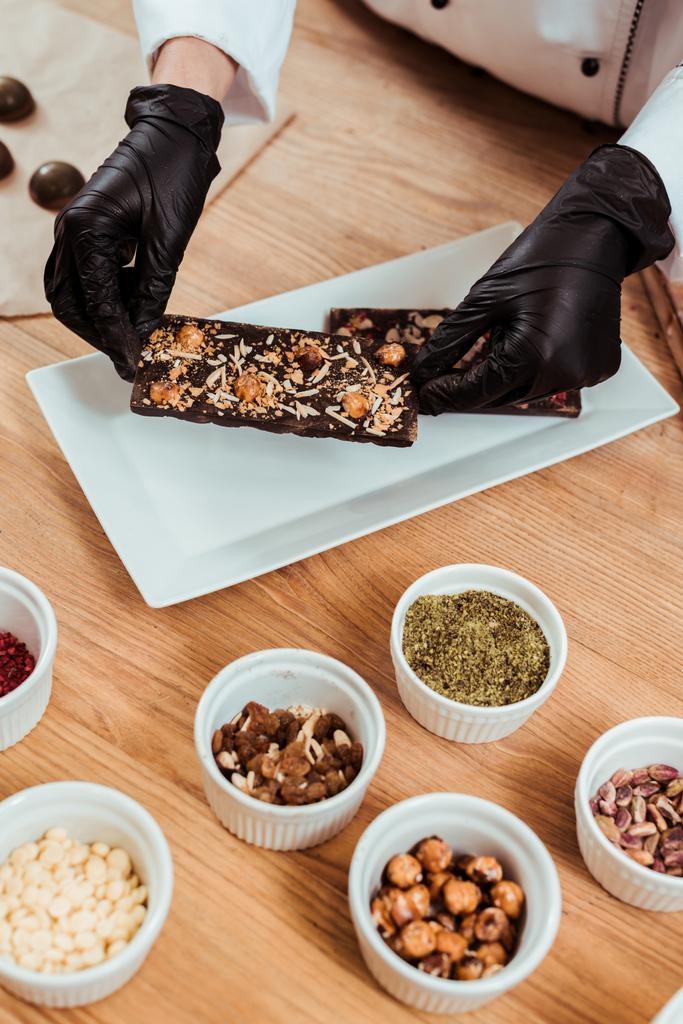 cropped άποψη της σοκολάτας σε latex γάντια βάζοντας σοκολάτα μπαρ με φουντούκια στο πιάτο  - Φωτογραφία, εικόνα