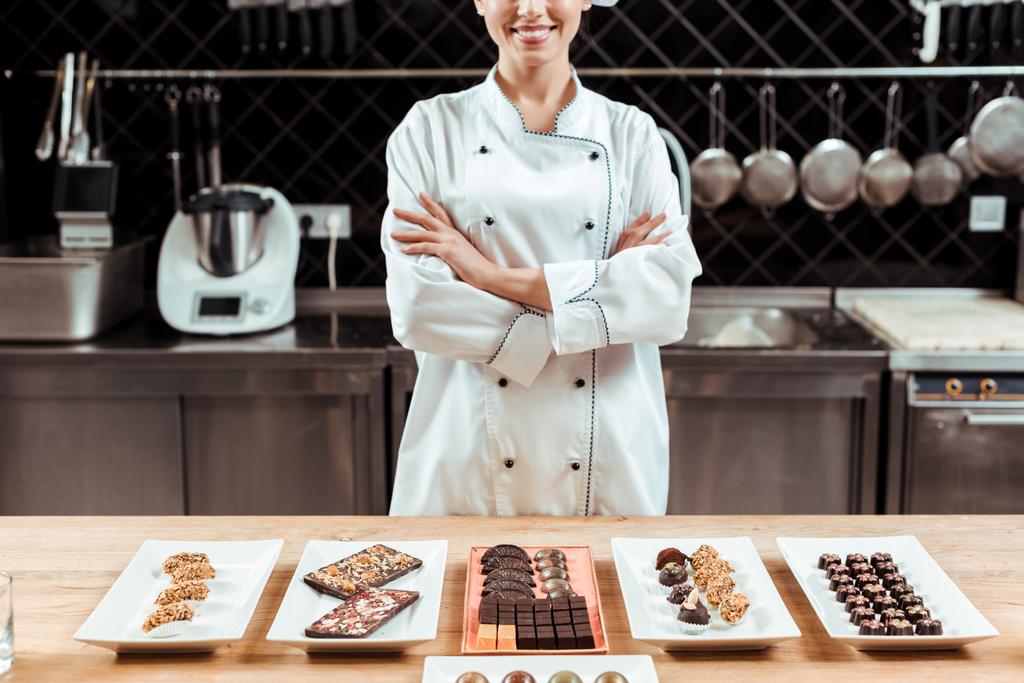 обрізаний вид щасливого шоколатьє, що стоїть з схрещеними руками біля смачних шоколадних цукерок на тарілках
  - Фото, зображення