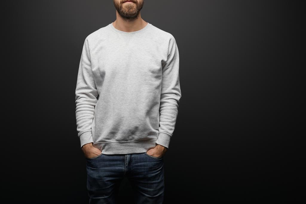 partial view of bearded man in blank basic grey sweatshirt with hands in pockets isolated on black - Photo, Image