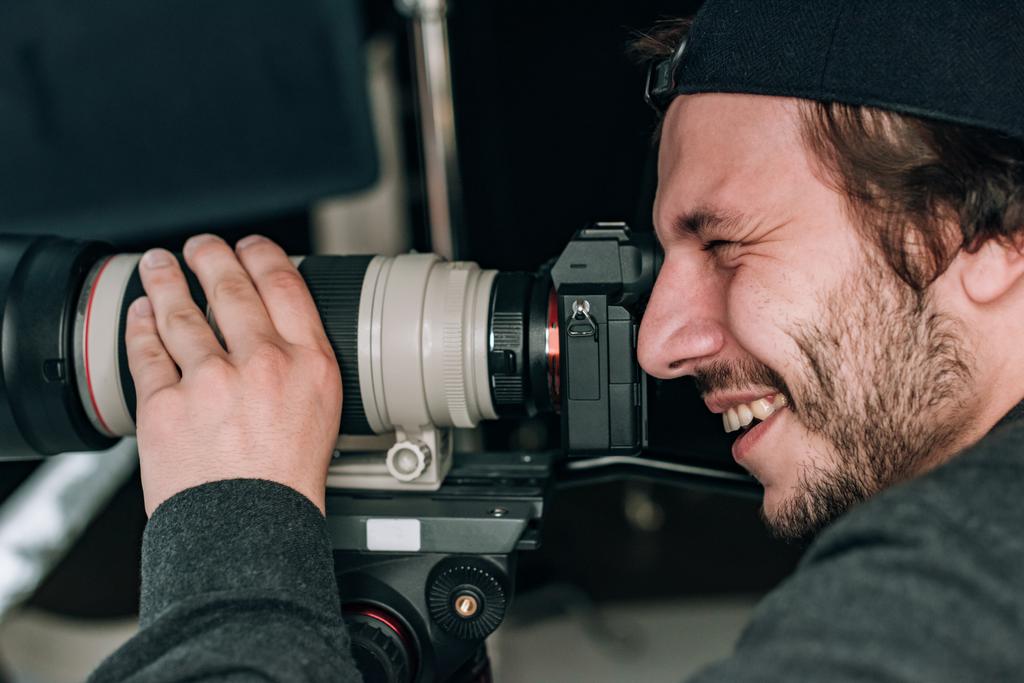 Zijaanzicht van lachende knappe videograaf met camera  - Foto, afbeelding