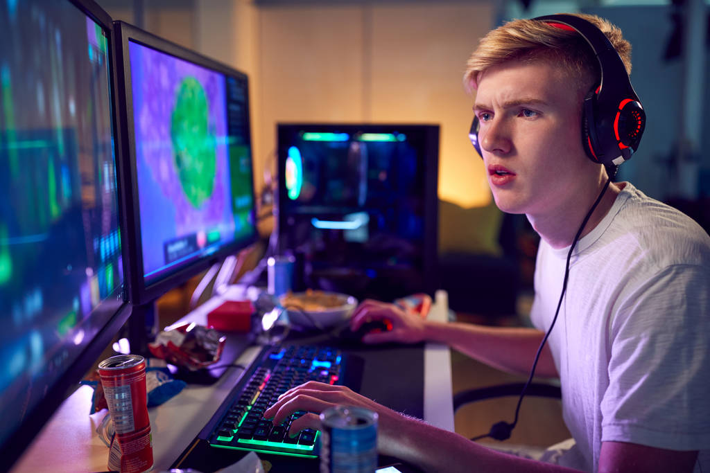 Teini-ikäinen poika yllään kuulokkeet Gaming kotona käyttämällä Dual tietokoneen näytöt
 - Valokuva, kuva
