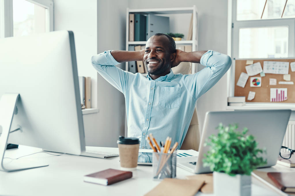 Hezký mladý Afričan v košili držet ruce za hlavou a usmívat se při práci v kanceláři     - Fotografie, Obrázek