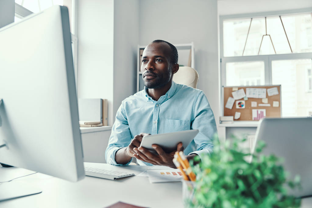 Задумчивый молодой африканский мужчина в рубашке с цифровым планшетом во время работы в офисе
                  - Фото, изображение