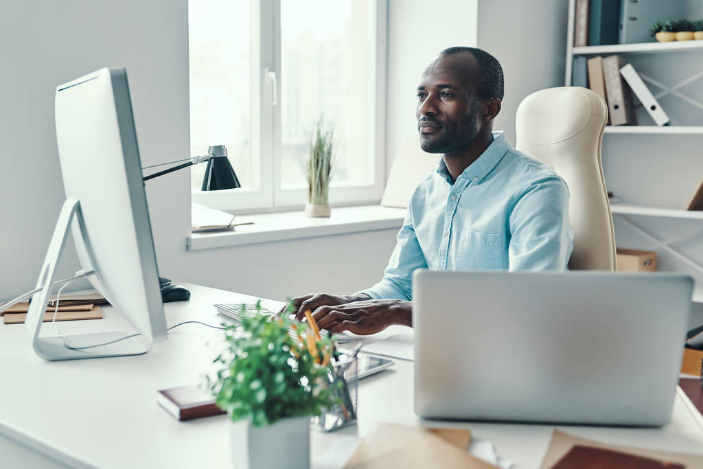 Koncentrált fiatal afrikai férfi ingben számítógépet használ munka közben az irodában        - Fotó, kép