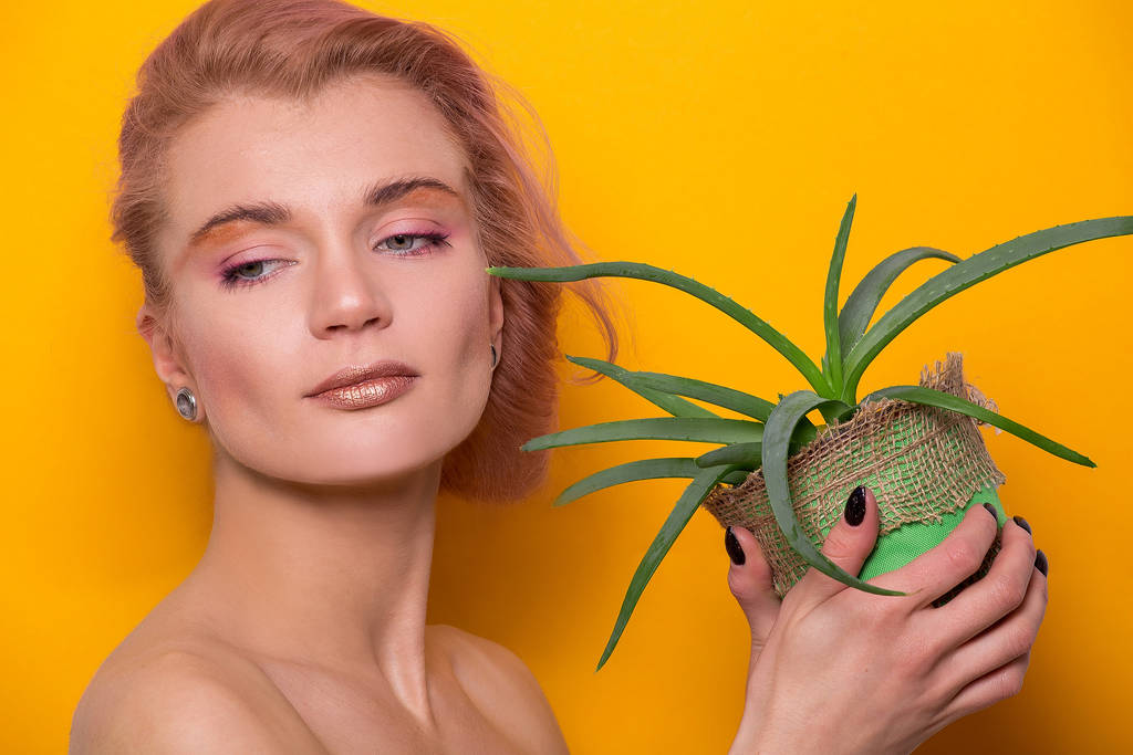 goldglänzendes Make-up für ein schönes junges europäisches Model, das in einem Fotostudio einen Aloe-Split in den Händen hält, isoliert auf orangefarbenem Hintergrund - Foto, Bild