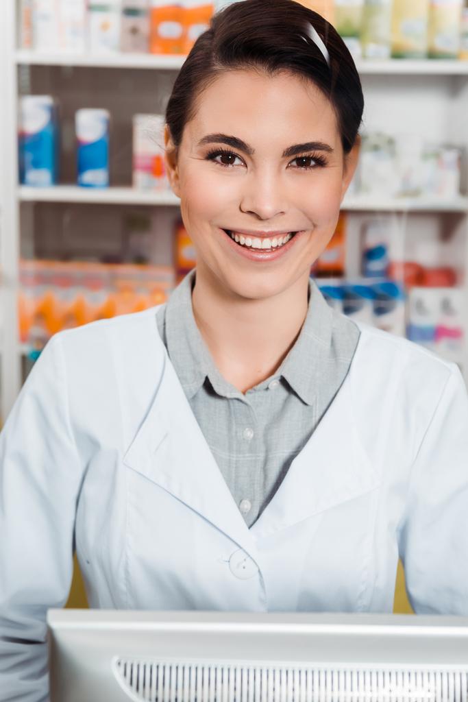 Привлекательный фармацевт улыбается в камеру с лекарствами на полках на заднем плане
 - Фото, изображение