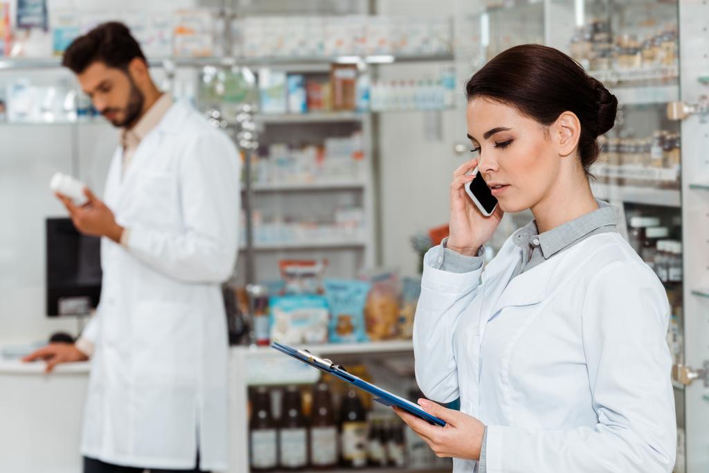 Szelektív fókusz a gyógyszerész vágólappal beszél okostelefonon kollégával a háttérben - Fotó, kép