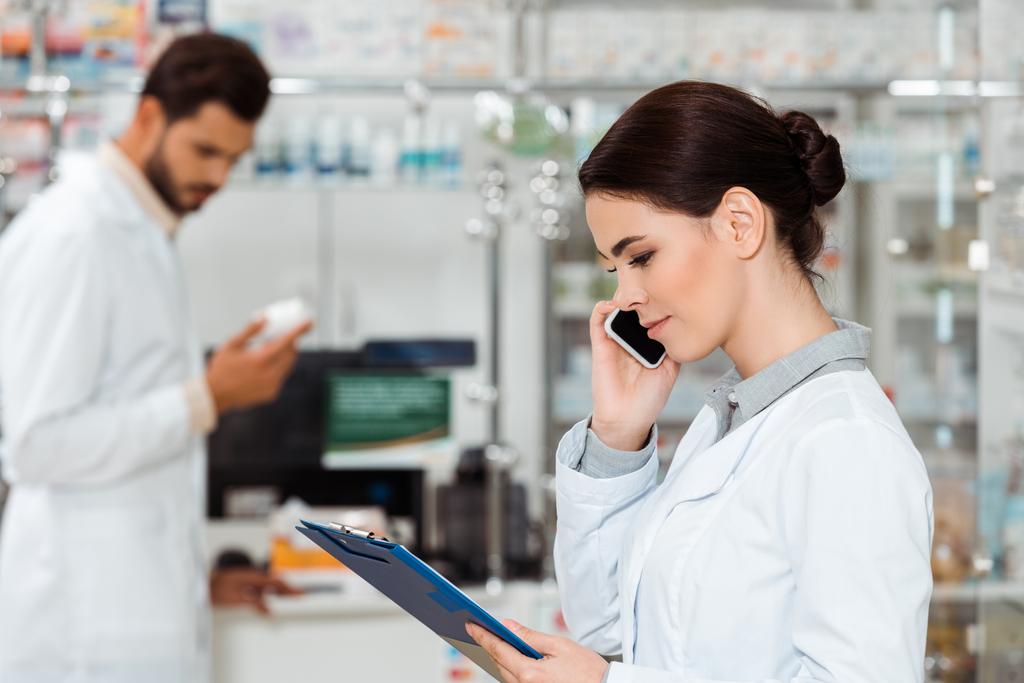 Selective focus of pharmacist with clipboard talking on smartphone with colleague holding pills at background - Photo, Image