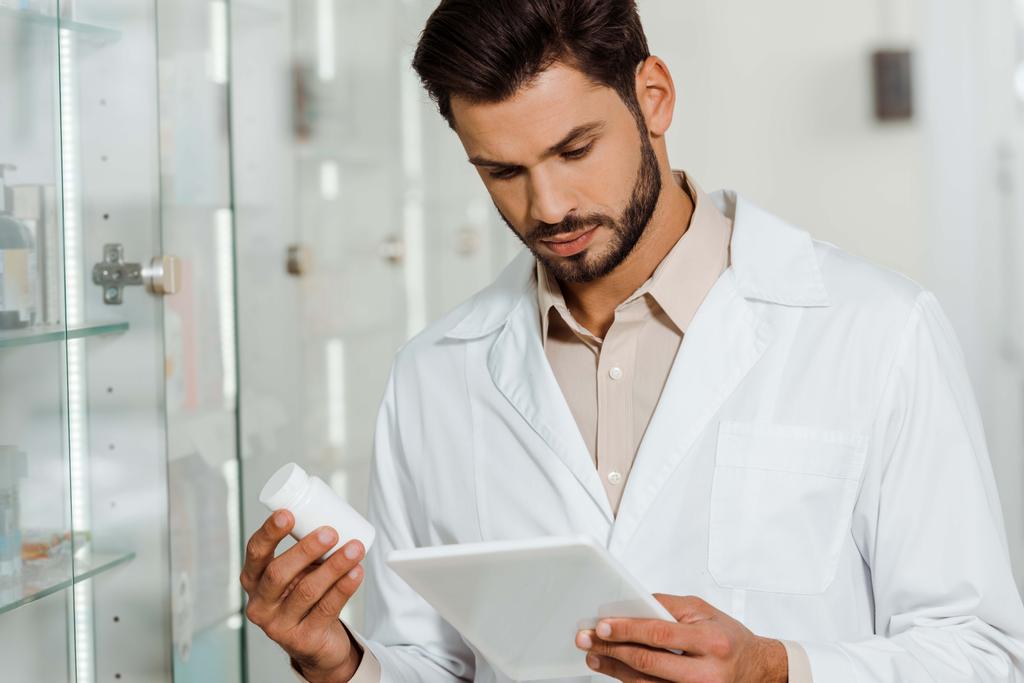 Guapo farmacéutico usando tableta digital mientras sostiene frasco de píldoras
  - Foto, Imagen